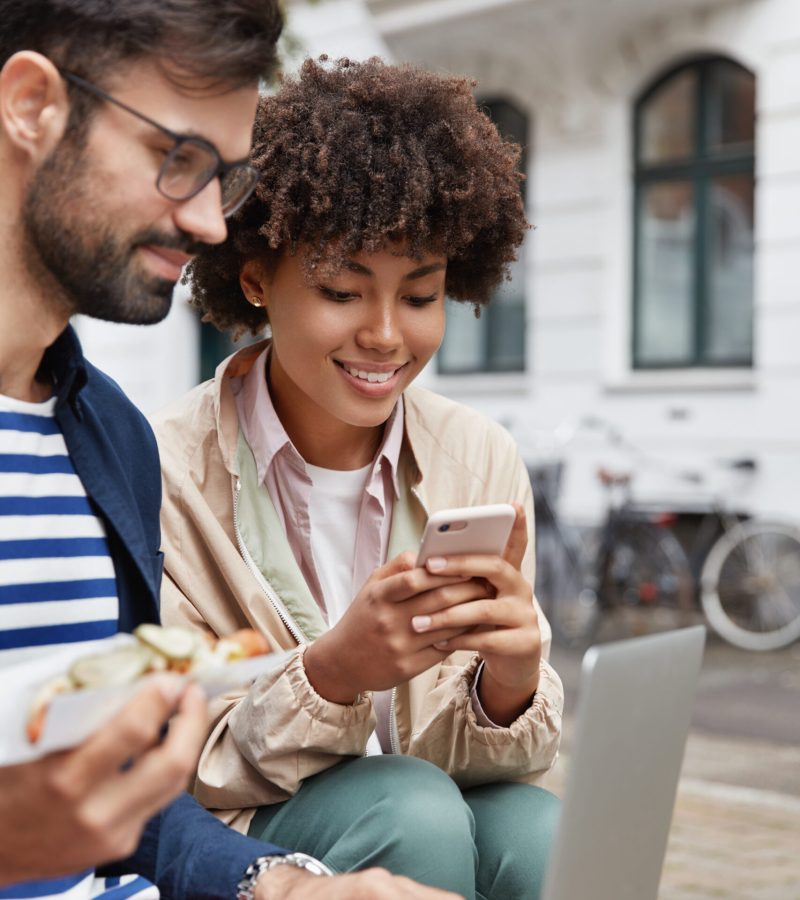 Youth addicted to modern technologies. Bearded hipster guy browses website for networking, eats junk food, attractive black woman with pleasant smile chooses item in web store, pose outdoor.